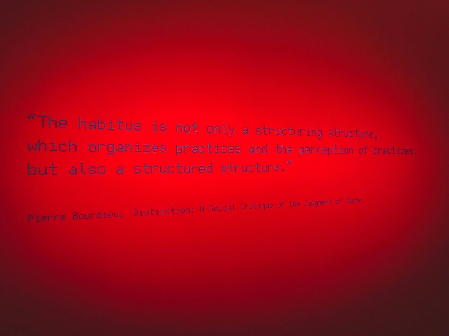 A red wall displays a quote by Pierre Bourdieu about habitus being both a structuring structure and a structured structure.