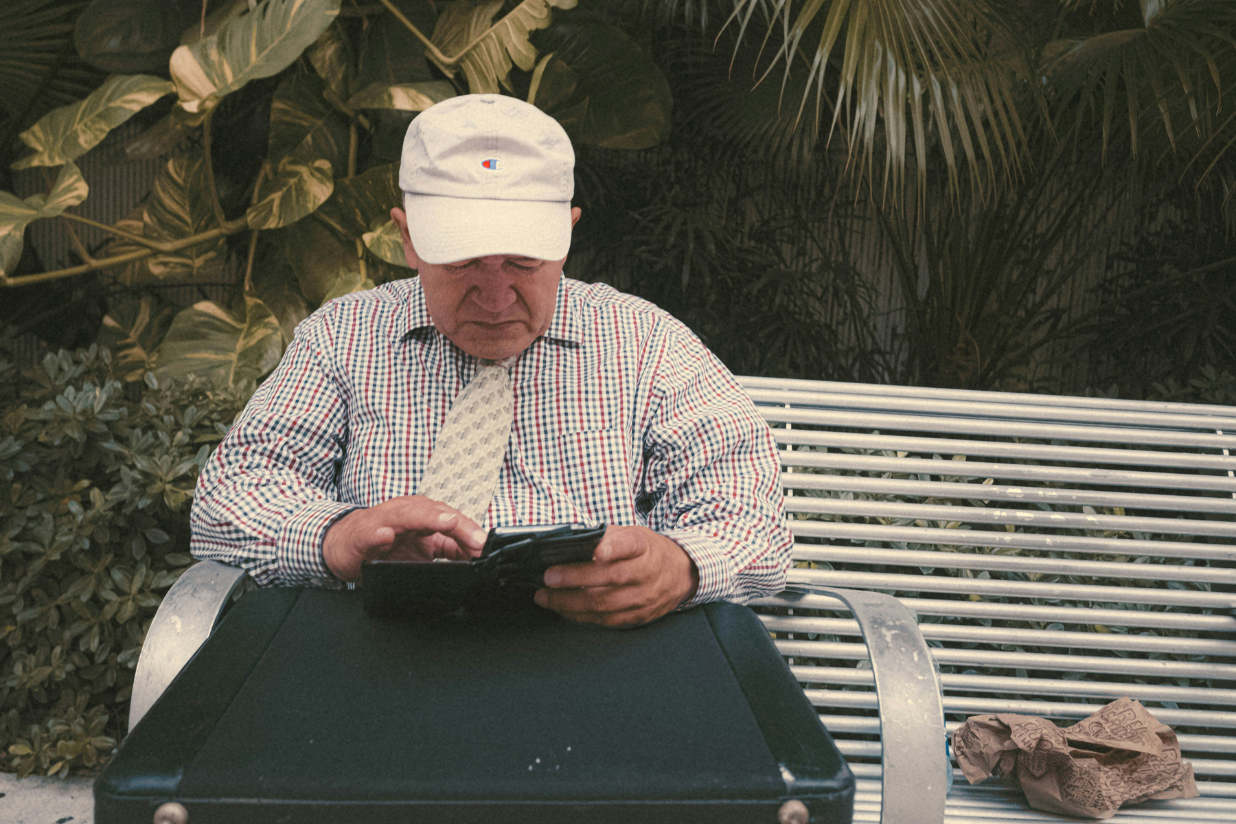 Auto-generated description: An older man wearing a cap and tie sits on a bench, focused on his phone or tablet, surrounded by lush greenery.