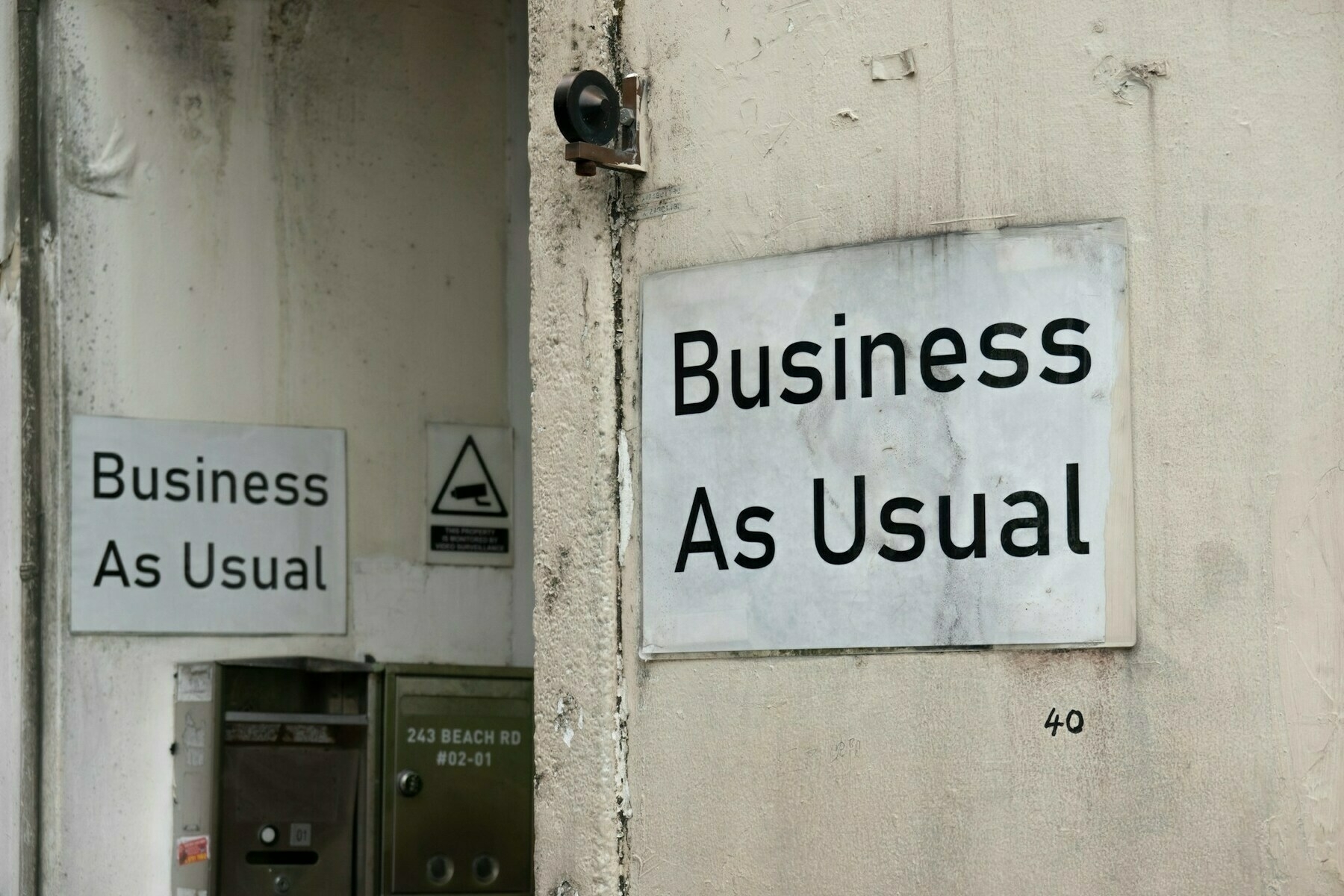Two signs stating Business As Usual are mounted on a weathered wall near a doorway.