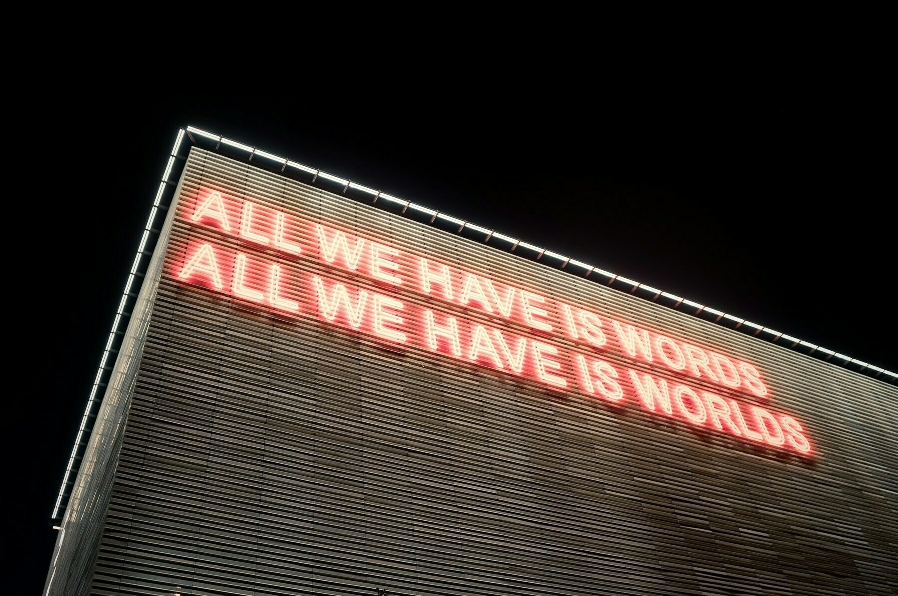 Auto-generated description: A large illuminated sign on a building reads ALL WE HAVE IS WORDS ALL WE HAVE IS WORLDS.