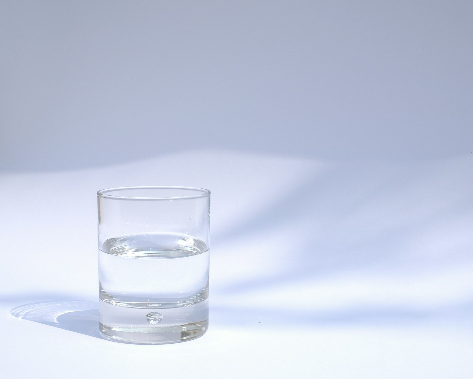Auto-generated description: A clear glass filled halfway with water casts a shadow on a white surface.
