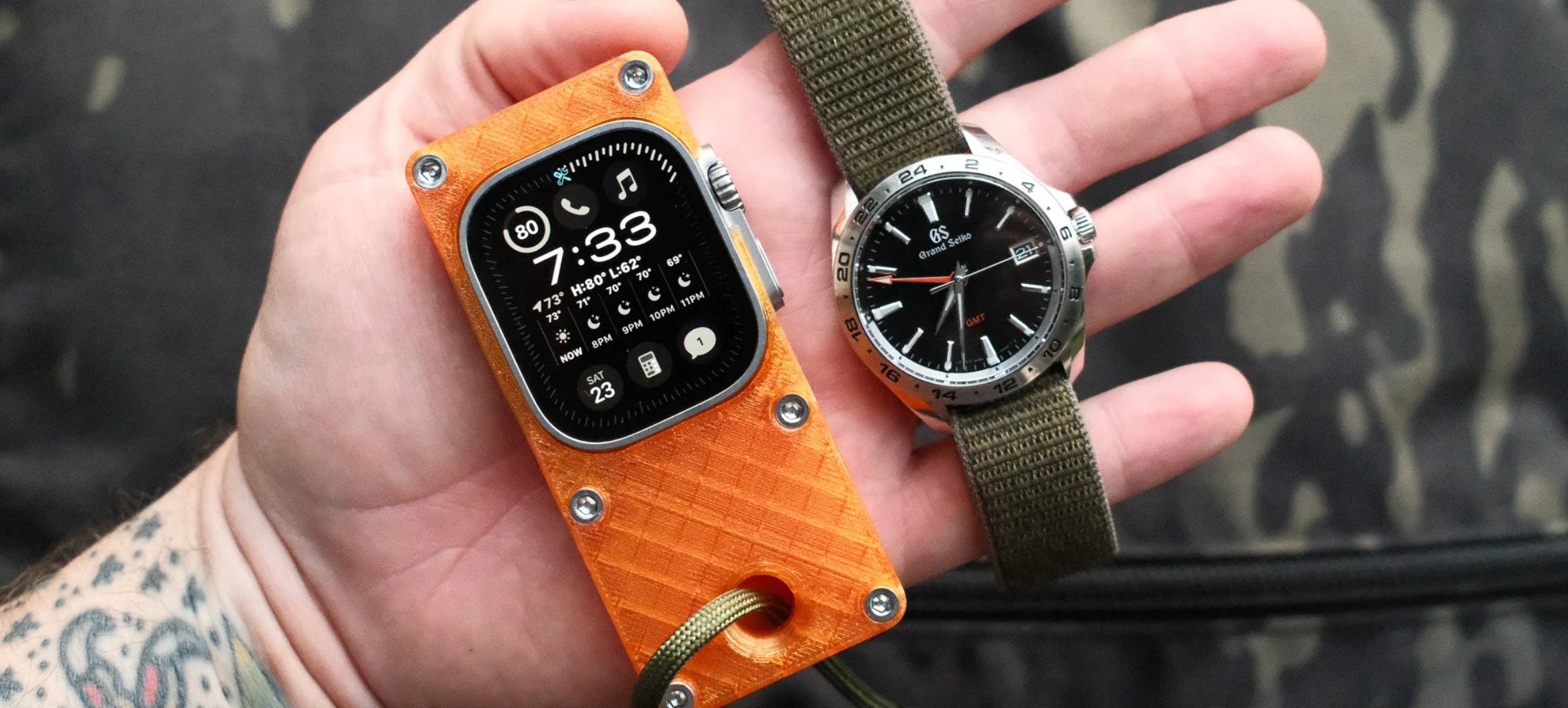Auto-generated description: A hand holds an Apple Watch in an orange case alongside an analog wristwatch with a dark dial and green strap.