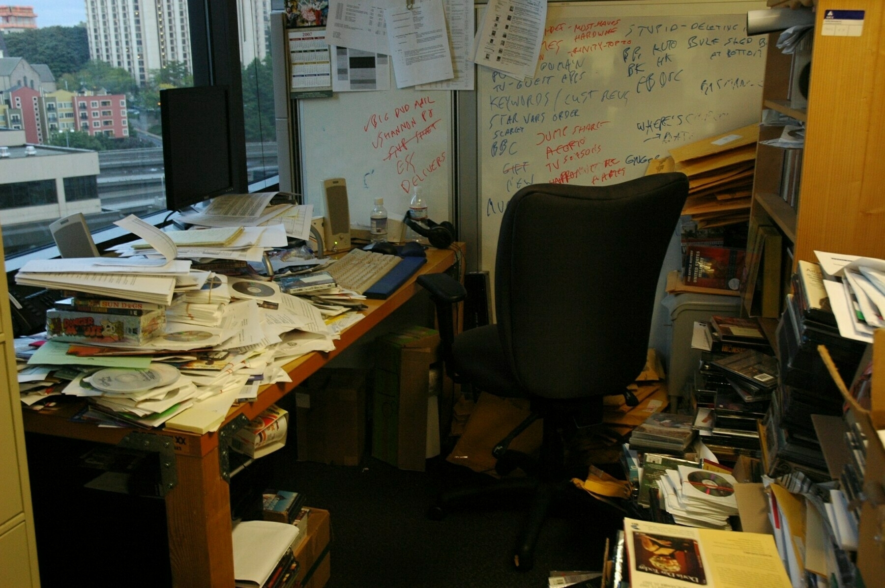 Auto-generated description: An office space is cluttered with piles of papers, books, and various items scattered around the desk and floor, with a chair positioned in front of a window.