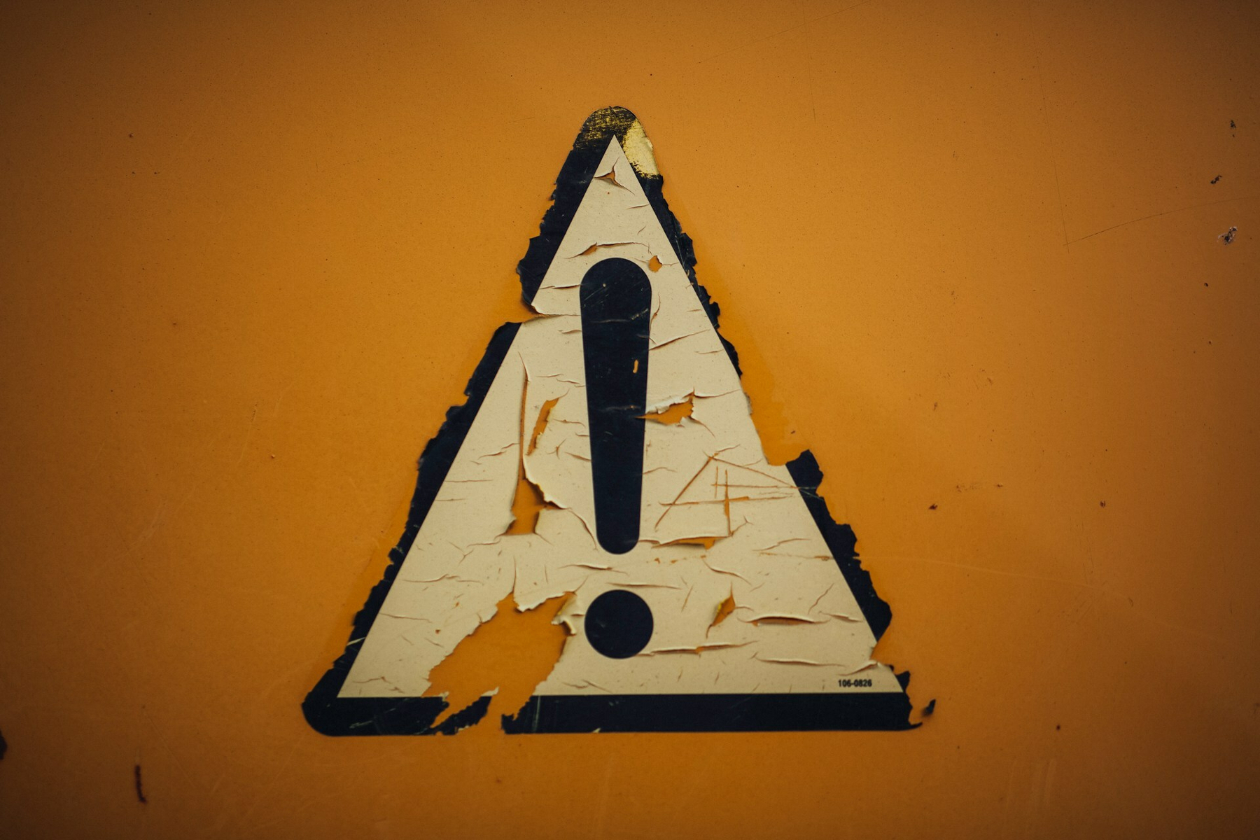 Auto-generated description: A weathered and peeling triangular warning sign with an exclamation mark is displayed against a yellow background.
