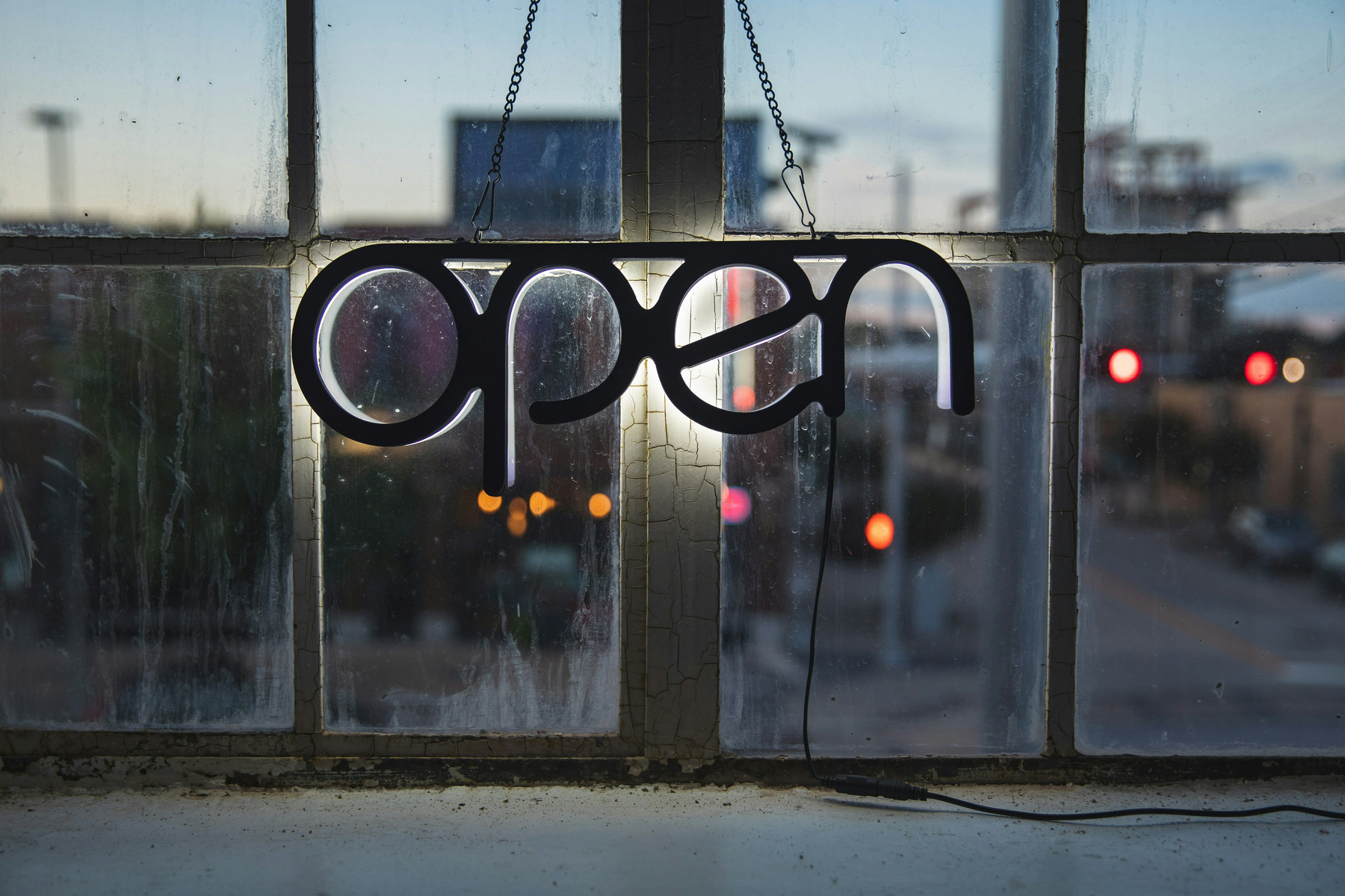 Auto-generated description: A neon open sign hangs in a window, with an urban street scene visible in the background at dusk.